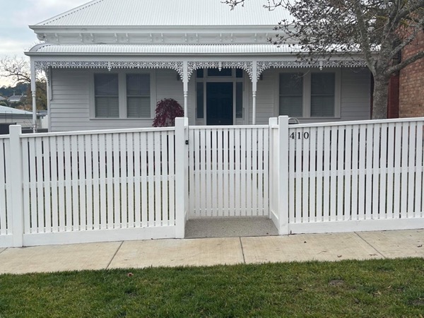 a white picket fence and gate in front of a weatherboard cottage